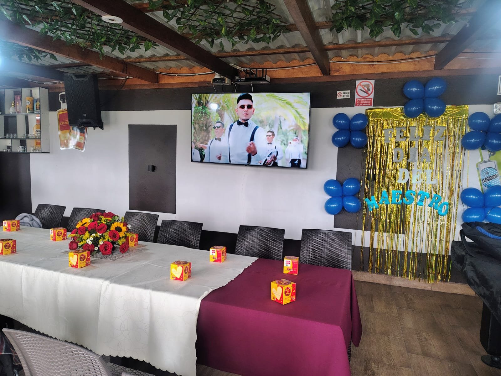Salón de eventos con mesas largas, globos azules y televisor.